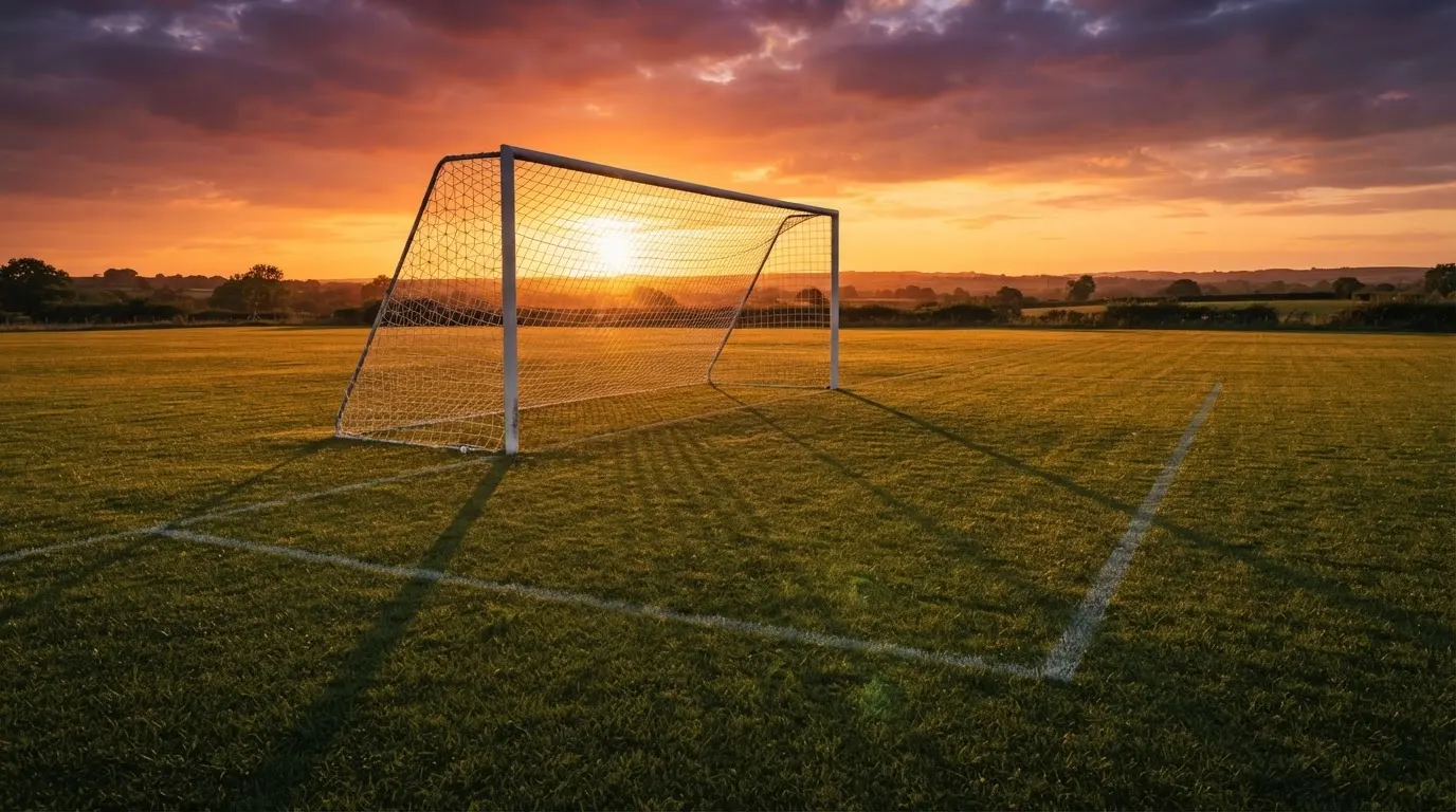 Ein Fußballtor mit Netz bei Sonnenuntergang auf einem leeren Platz, die Schatten der Torpfosten liegen lang auf dem Gras.