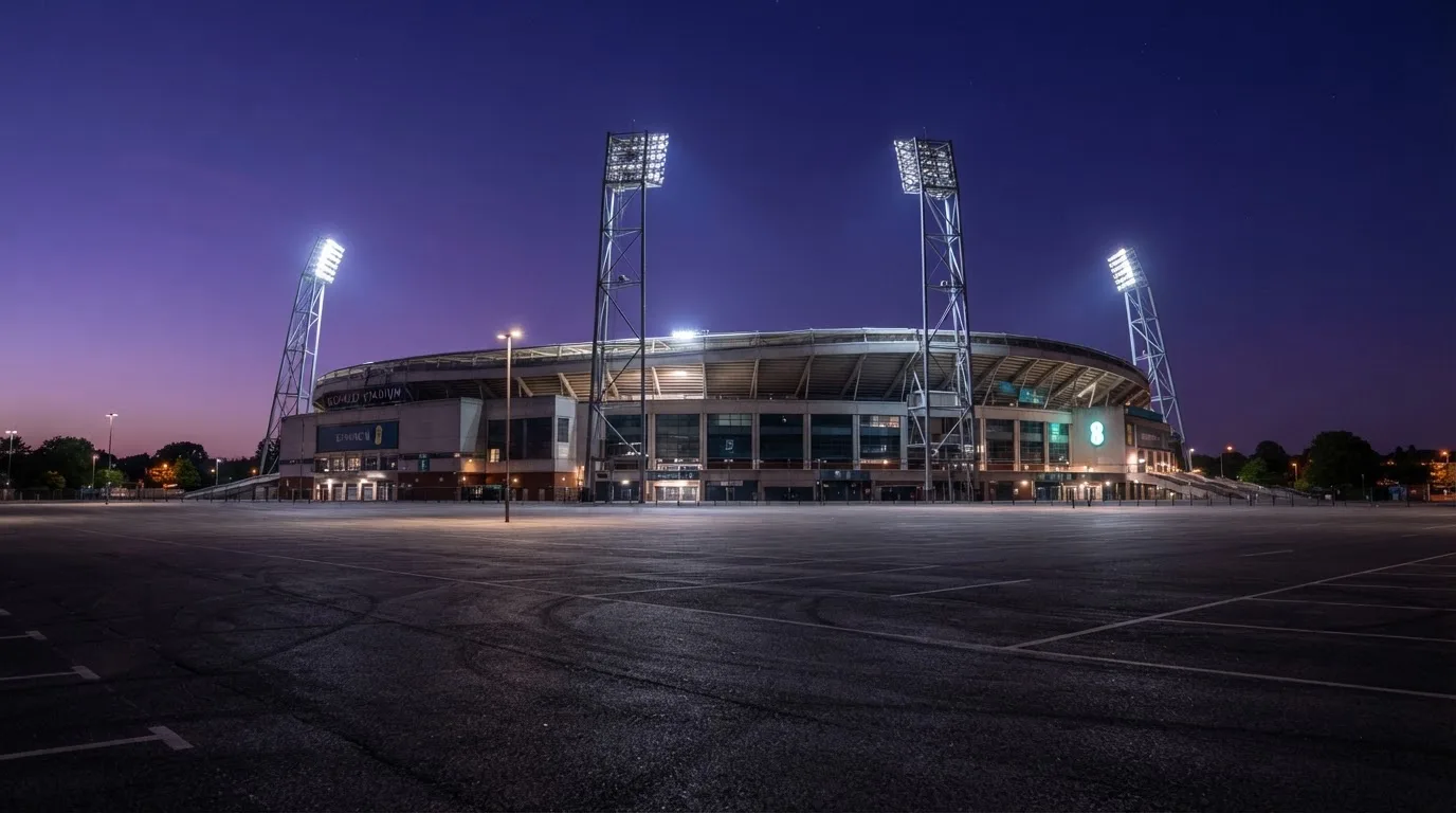 Ein Fußballstadion von außen in der Dämmerung, hohe Flutlichtmasten vor einem violetten Himmel, davor ein leerer Parkplatz.