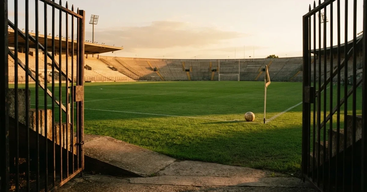 Blick durch ein Stadiontor auf einen grünen Rasen im Abendlicht, ein Ball ruht an der Eckfahne, die Tribünen sind leer.
