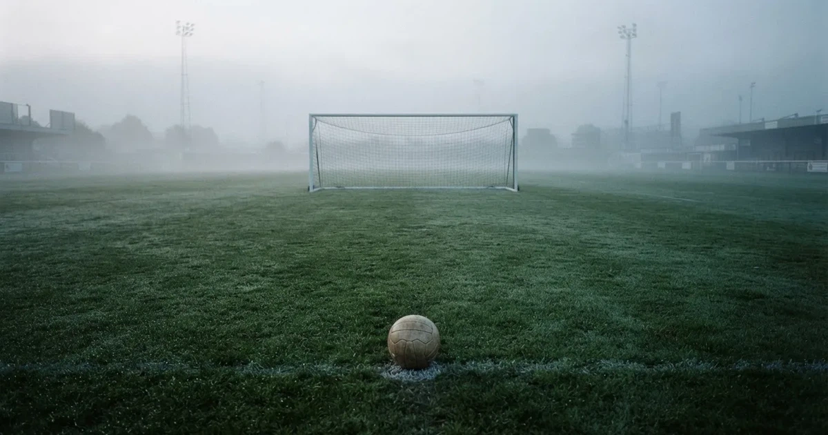 Ein Fußballtor mit gespanntem Netz steht auf einem leeren Rasenplatz, davor ein einzelner Ball, Morgendunst liegt über dem Feld.