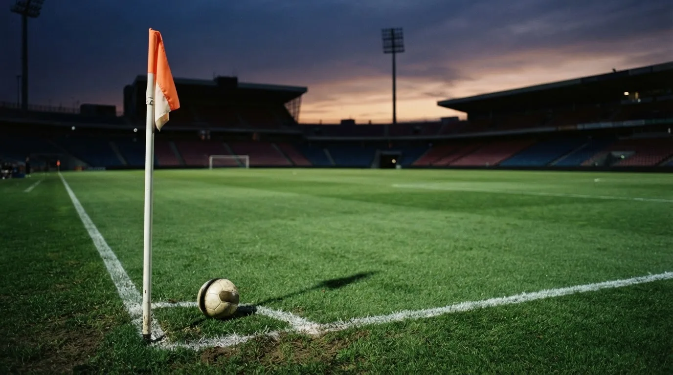Ein Fußball liegt neben einem Eckfahnen-Pfosten auf einem Rasenplatz, im Hintergrund ein leeres Stadion in der Dämmerung.
