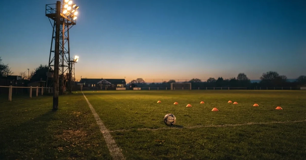 Eine Flutlichtanlage ragt über einen Trainingsplatz, auf dem Rasen liegen verstreut Hütchen und ein Ball, am Himmel die Abenddämmerung.