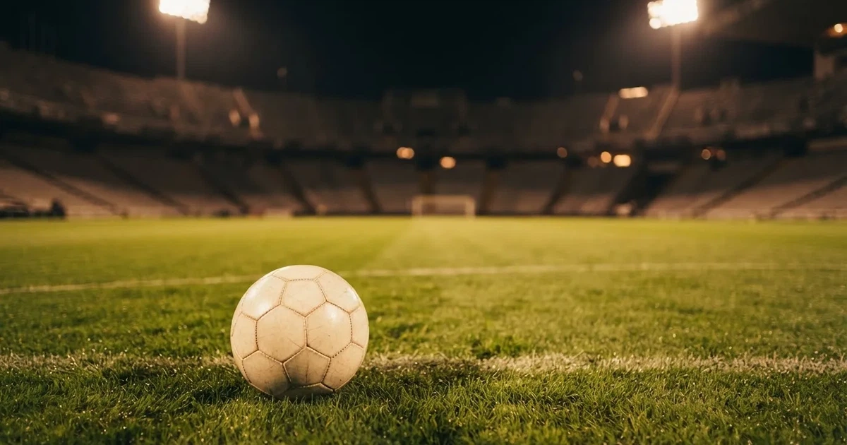 Ein Fußball liegt auf dem Rasen eines leeren Stadions bei Flutlicht, im Hintergrund unscharfe Tribünen ohne erkennbare Beschriftung.