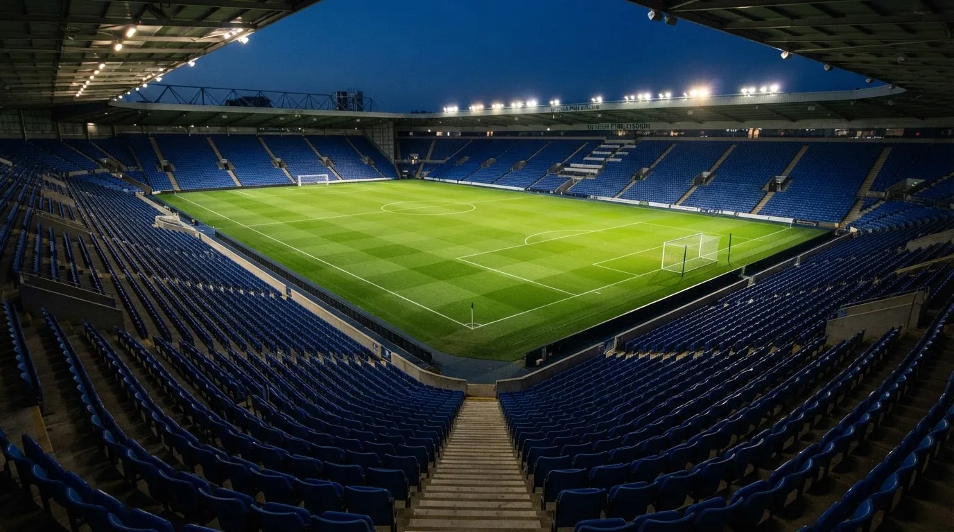 Eine leere Stadiontribüne unter Flutlicht aus der Ferne betrachtet, die Rasenfläche leuchtet grün in der Dämmerung.