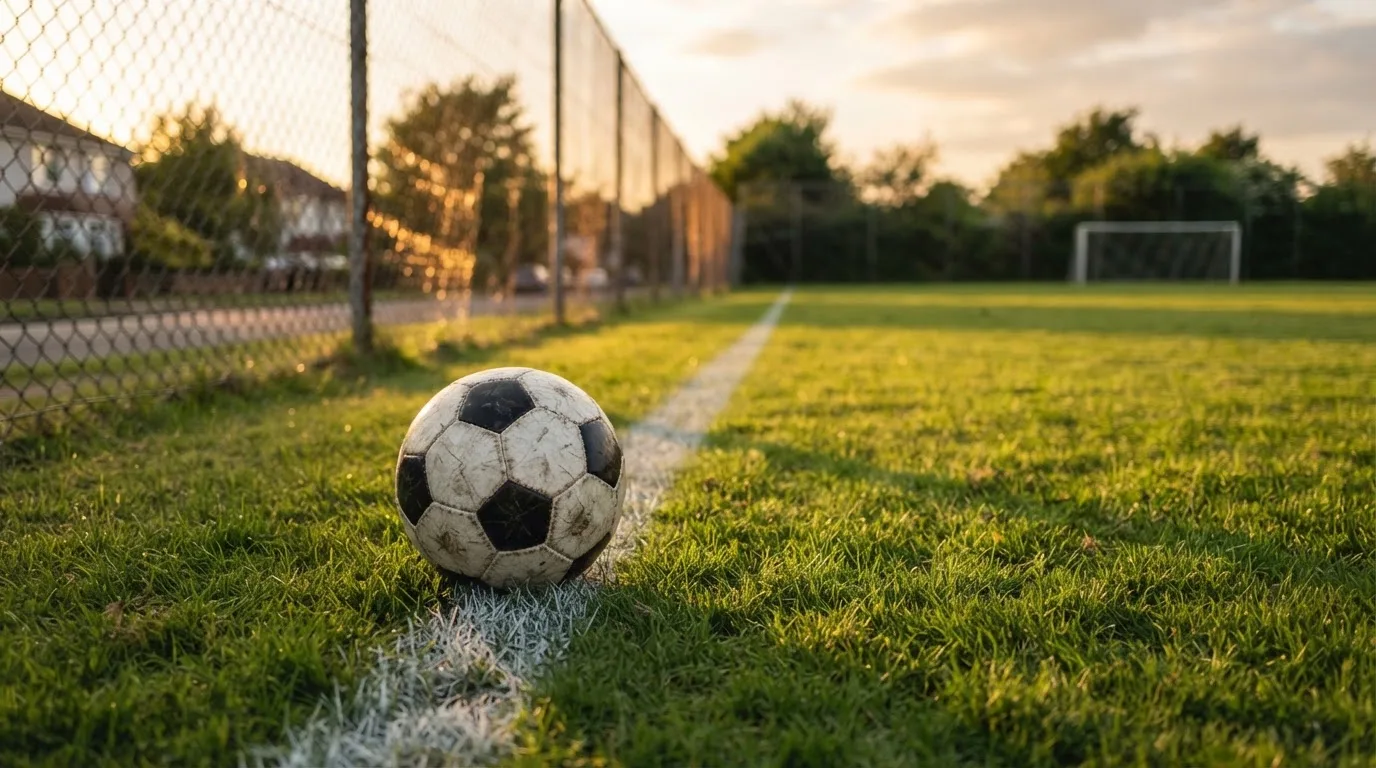 Ein Fußball liegt am Rand eines Rasens neben einer weißen Linie, im Hintergrund ein Zaun in der Abendsonne.