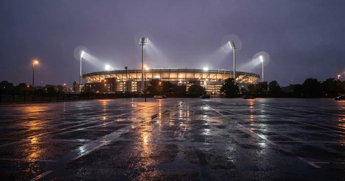 Ein Stadion unter Flutlicht aus großer Entfernung gesehen, davor ein regennasser Parkplatz in der Dämmerung.