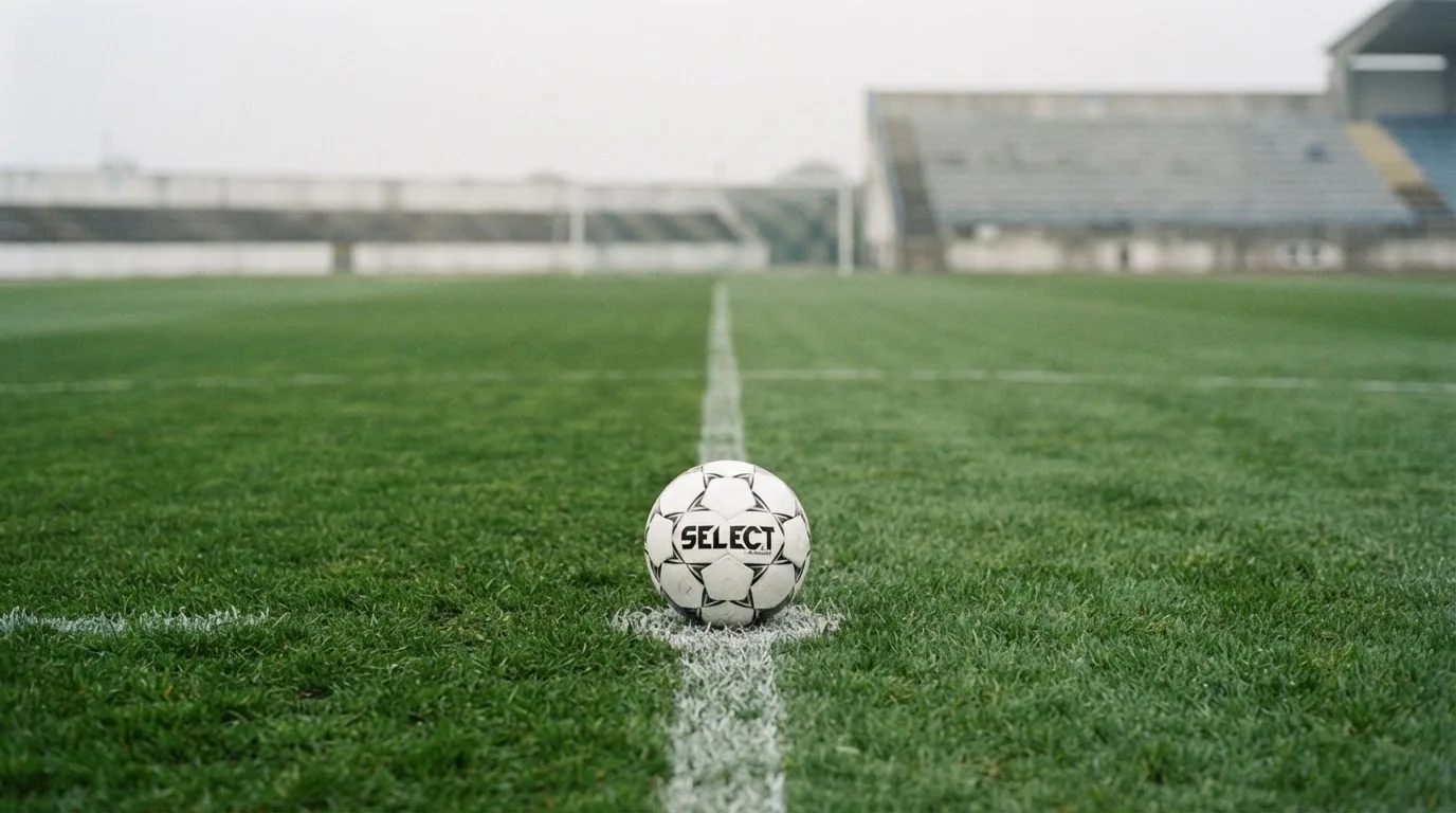 Ein Fußball liegt auf dem Anstoßpunkt eines leeren Rasenplatzes, im Hintergrund eine ruhige Tribüne unter bedecktem Himmel.