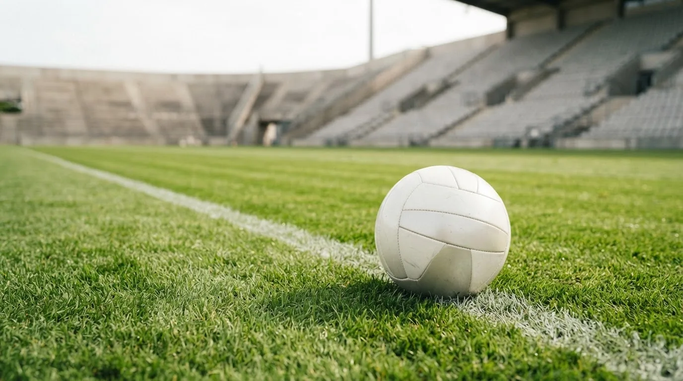 Ein Fußball liegt am Spielfeldrand eines Rasenplatzes, im Hintergrund eine leere Tribüne unter hellem Himmel.