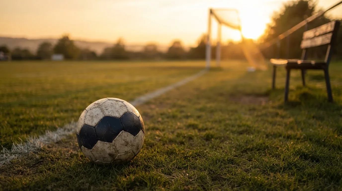 Ein Fußball liegt im Gras am Spielfeldrand, dahinter eine leere Bank und ein Tor im Gegenlicht.