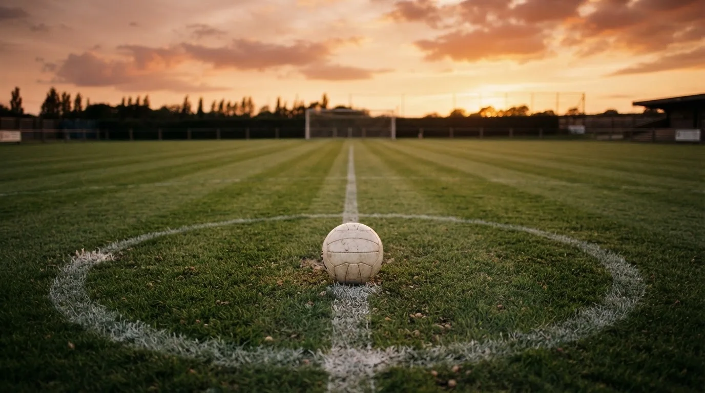 Ein Fußball liegt in der Mitte eines leeren Rasenplatzes, am Horizont färbt sich der Himmel orange.