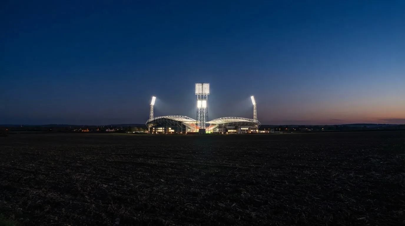 Ein Stadion unter Flutlicht aus großer Entfernung, davor ein dunkler Acker im Abendlicht.