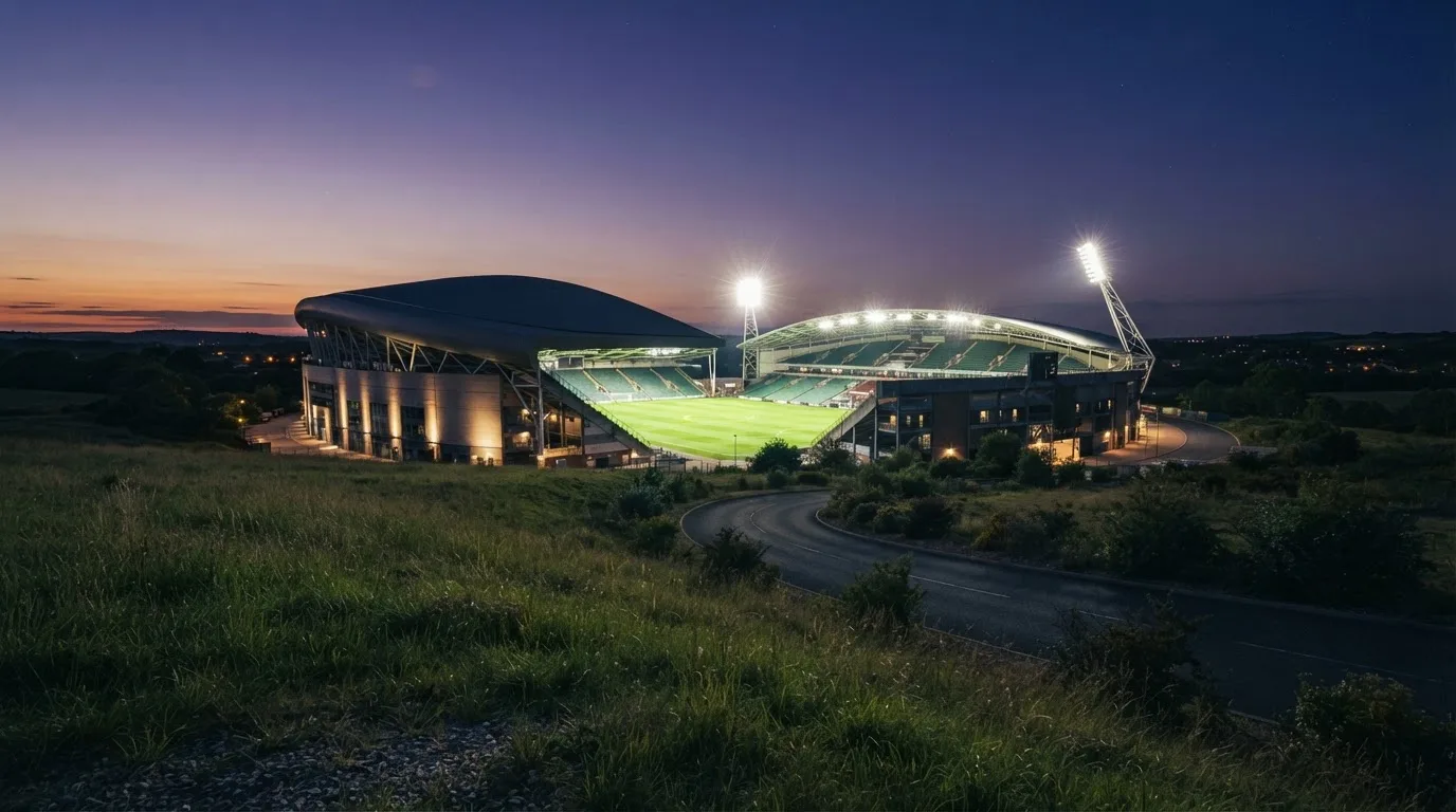 Ein Stadion unter Flutlicht, von einer Anhöhe aus gesehen, davor ein stiller Weg in der Dämmerung.