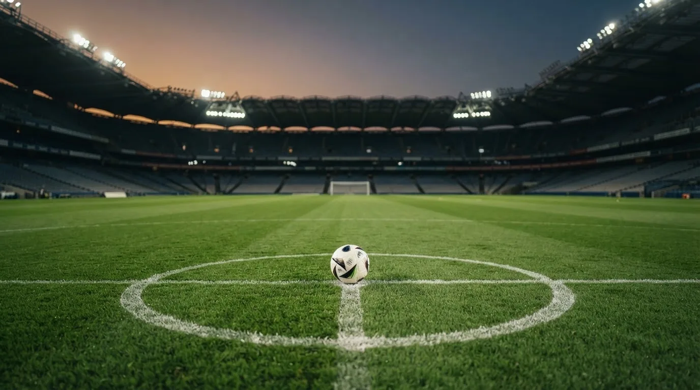 Ein Fußball liegt auf dem Anstoßpunkt eines leeren Rasenplatzes, im Hintergrund ein Stadion unter Flutlicht in der Abenddämmerung.