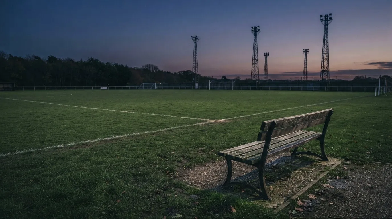 Eine leere Parkbank am Rand eines Fußballfelds in der Abenddämmerung, im Hintergrund ragen dunkle Flutlichtmasten in den Himmel.