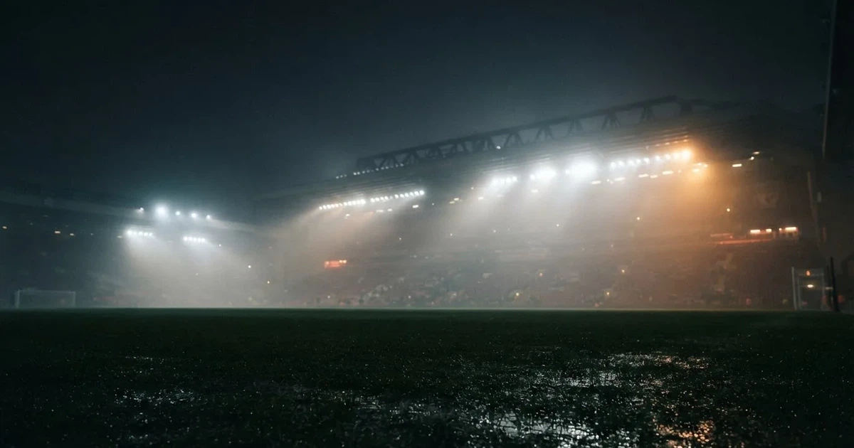 Ein Stadion unter Flutlicht, aus großer Entfernung über eine dunkle Wiese fotografiert, am Himmel zieht Nebel auf.