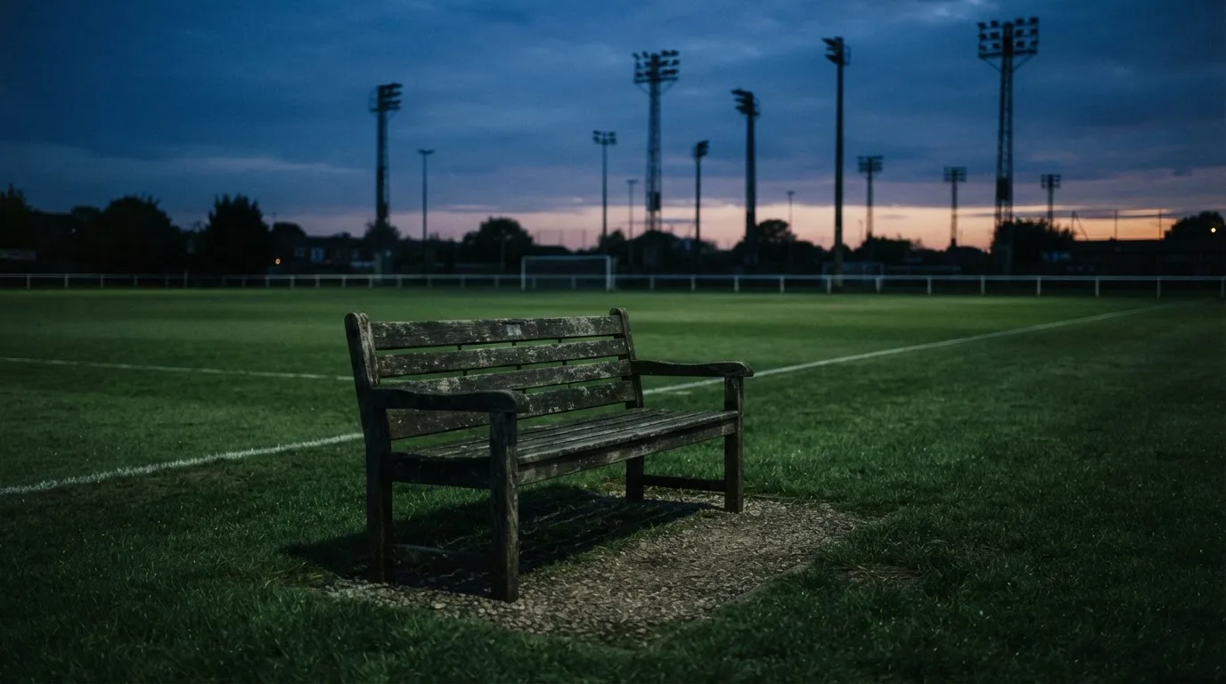 Eine leere Parkbank am Rand eines Fußballfelds in der Abenddämmerung, im Hintergrund ragen dunkle Flutlichtmasten in den Himmel.