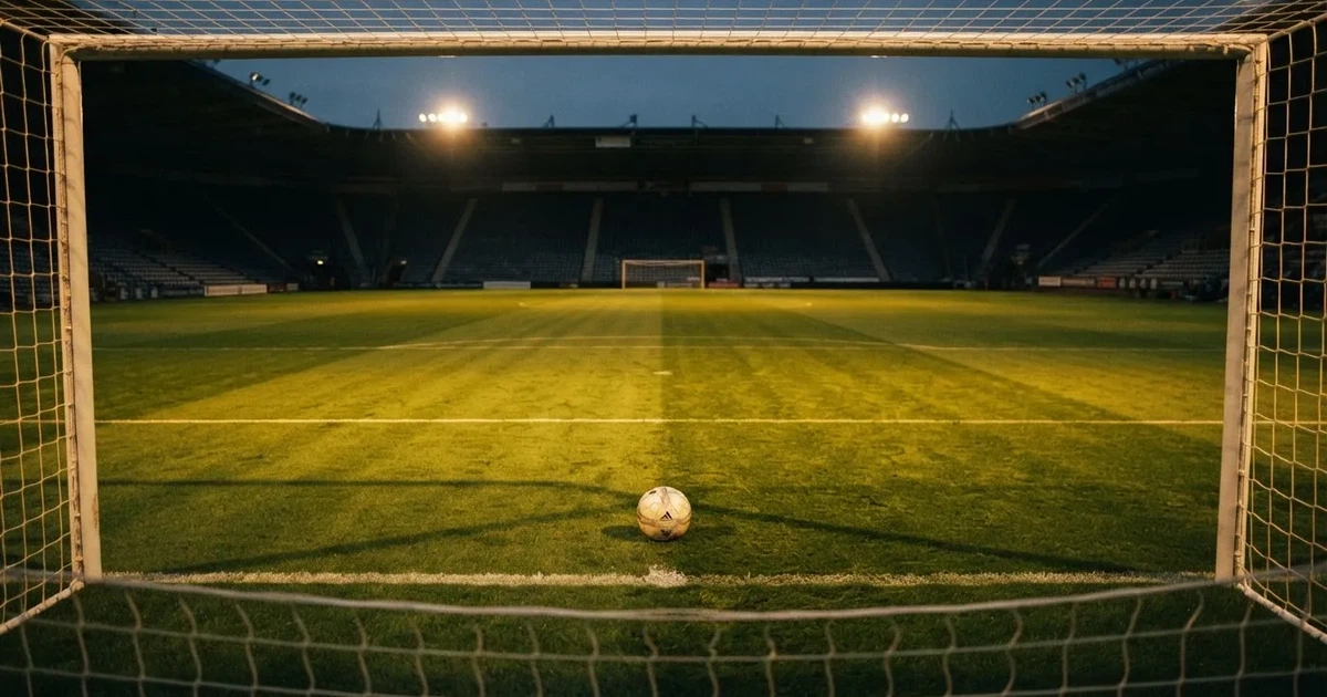 Ein Fußballplatz mit frisch gemähtem Rasen im Abendlicht, ein Ball liegt im Strafraum, im Hintergrund unscharf eine leere Tribüne.
