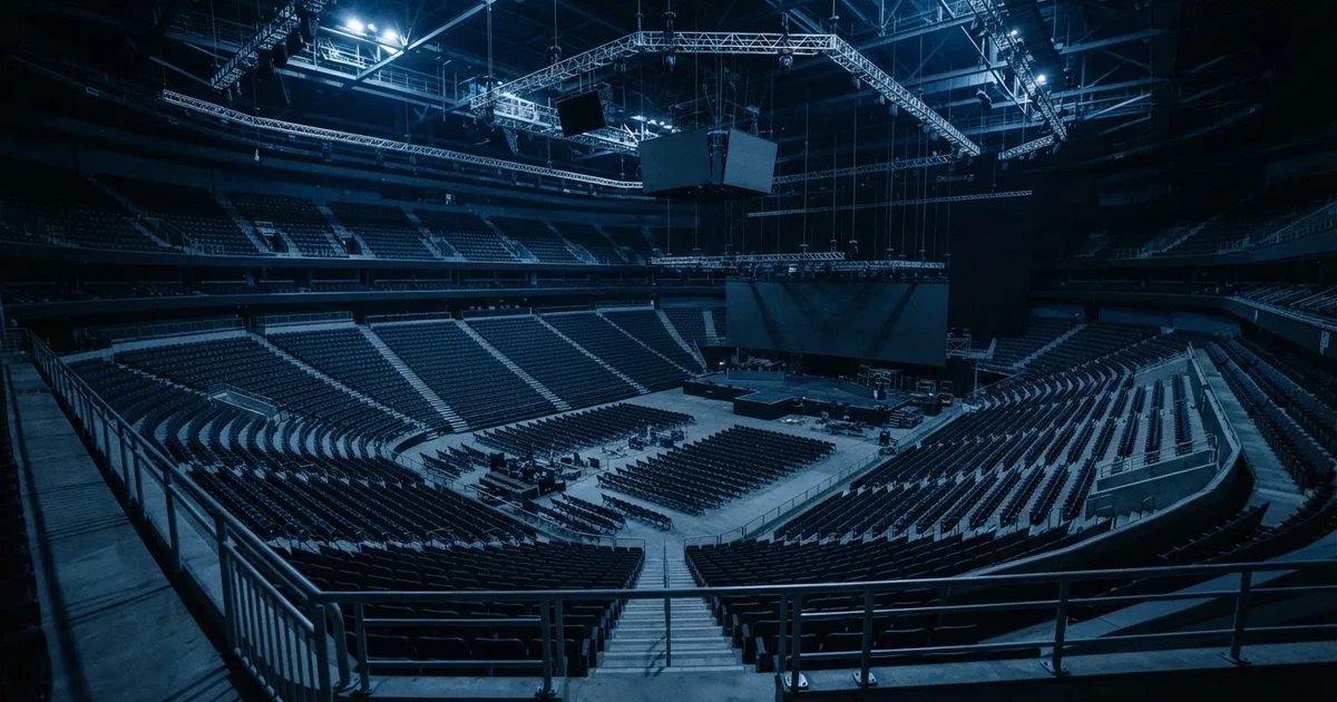 Eine große leere Arena mit Bühne und Stuhlreihen im Halbdunkel, von einem oberen Rang aus gesehen, alle Bildschirme ausgeschaltet.