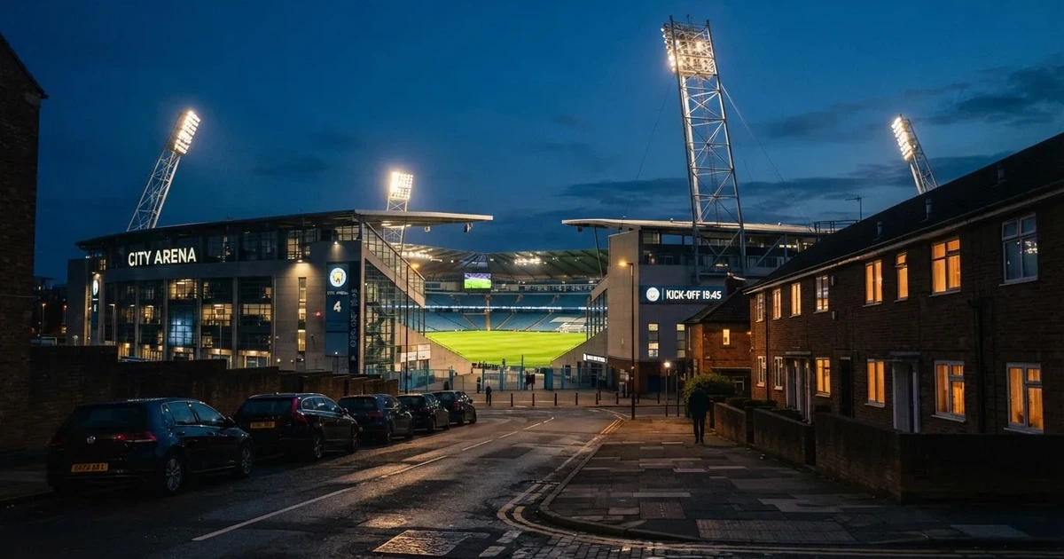 Ein Fußballstadion bei Flutlicht aus der Ferne gesehen, davor eine Straße und leuchtende Fenster eines Nebengebäudes.