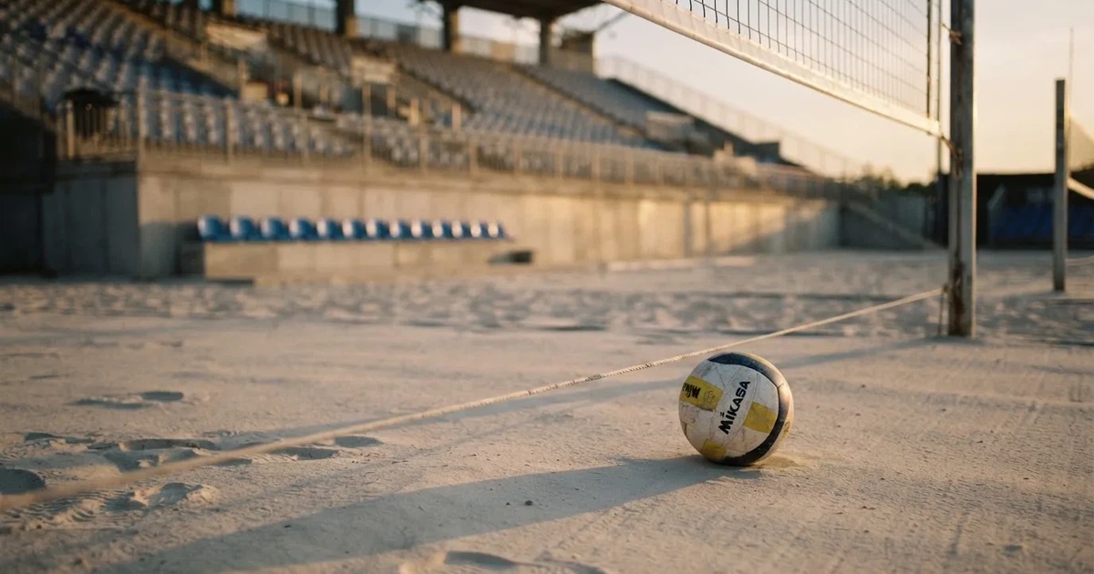 Ein Volleyball liegt auf dem Sand eines Beachvolleyball-Feldes neben dem gespannten Netz, im Hintergrund eine leere Tribüne bei Abendlicht.