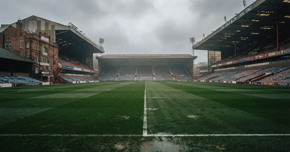 Ein englisches Fußballstadion mit rotbraunen Backsteintribünen unter bedecktem Himmel, davor ein nasser Rasen mit Spielfeldlinien.