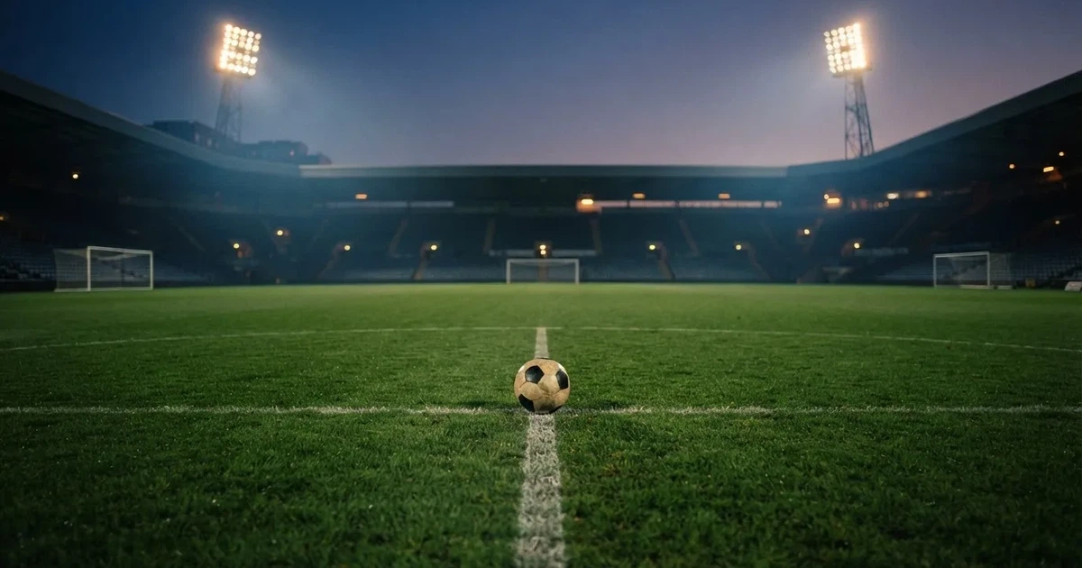 Ein Rasenplatz in der Dämmerung mit eingeschalteten Flutlichtmasten, ein Ball liegt am Anstoßpunkt, die Tribünen im Hintergrund sind unscharf.