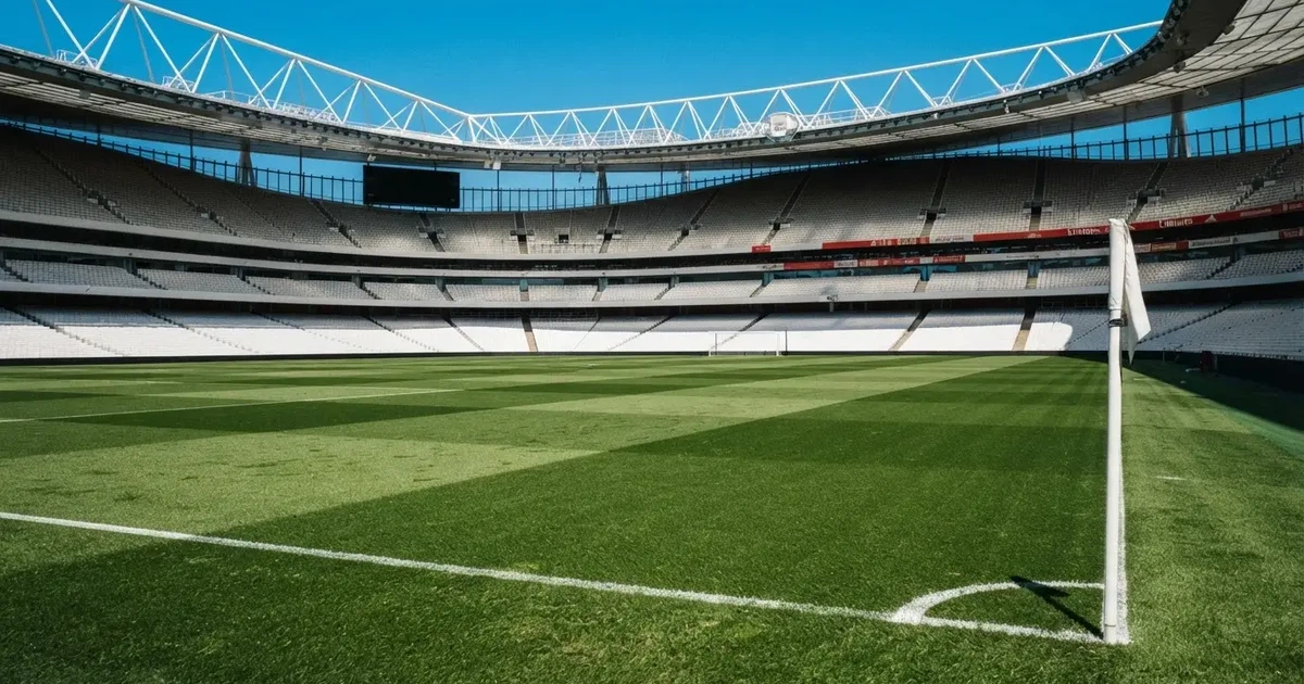 Ein Fußballstadion mit hellen Rängen unter strahlend blauem Himmel, davor ein sonnenbeschienener Rasen mit frisch gemähten Streifen.