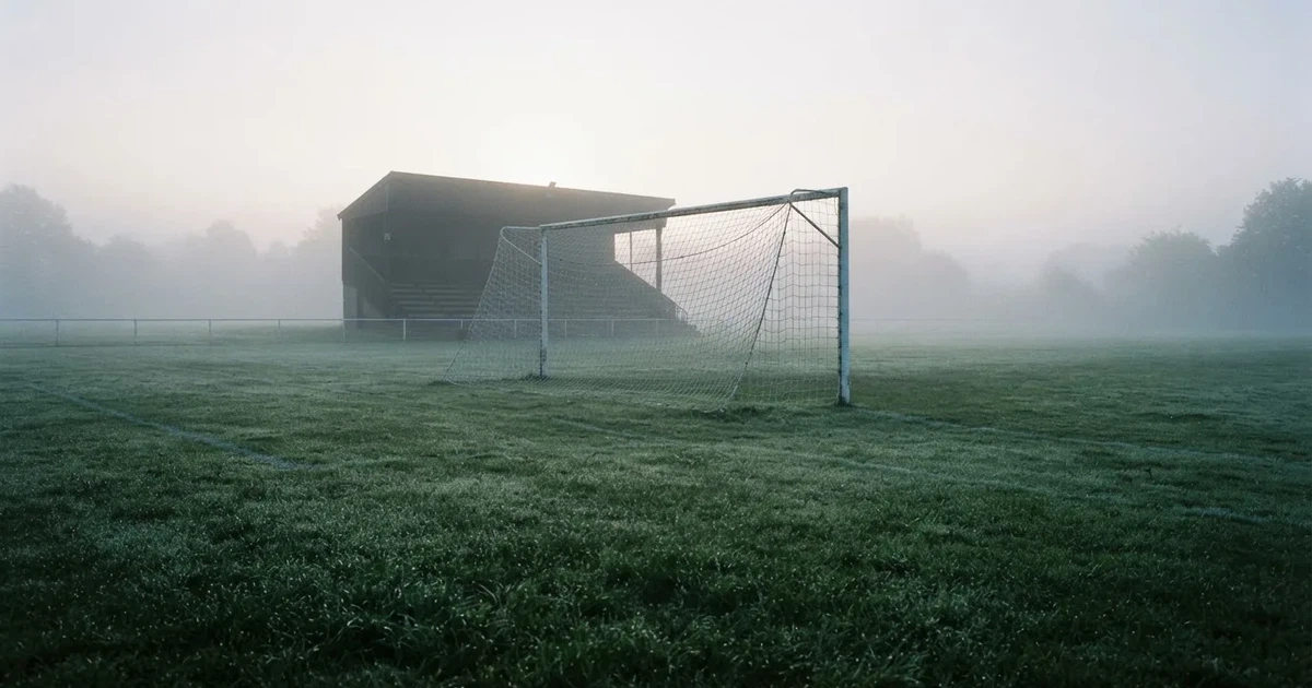 Ein Fußballtor mit Netz steht im Morgennebel auf einem leeren Rasenplatz, im Hintergrund die Silhouette einer kleinen Tribüne.