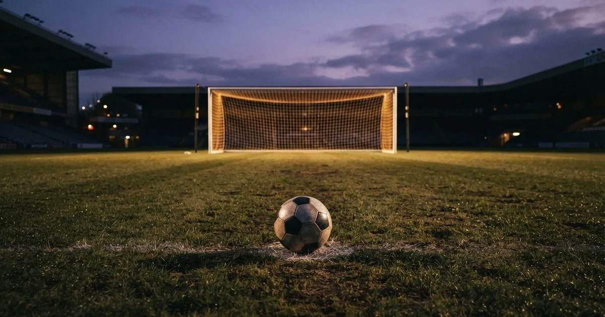 Ein Fußballtor mit Netz im Gegenlicht auf einem leeren Platz am Abend, davor ein Ball auf dem Rasen.