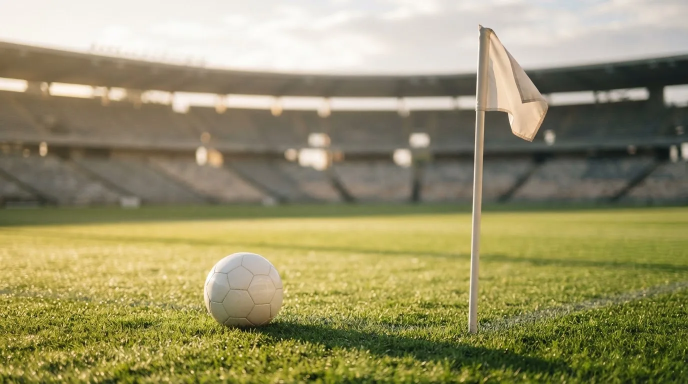 Ein Fußball liegt auf Rasen neben einer Eckfahne ohne Aufdruck auf einem leeren Platz im Morgenlicht.
