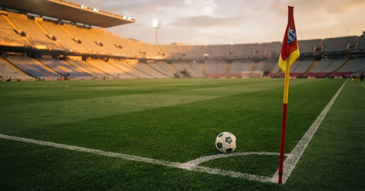 Ein Fußball liegt am Viertelkreis an der Eckfahne eines Rasenplatzes, im Hintergrund leere Tribünen in warmem Abendlicht.