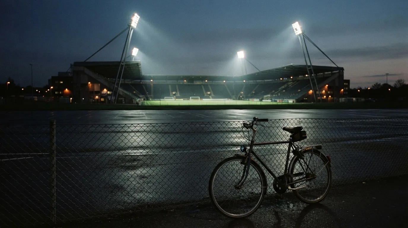 Ein Stadion unter Flutlicht aus der Ferne gesehen, davor ein dunkler Parkplatz und ein einzelnes Fahrrad an einem Zaun.