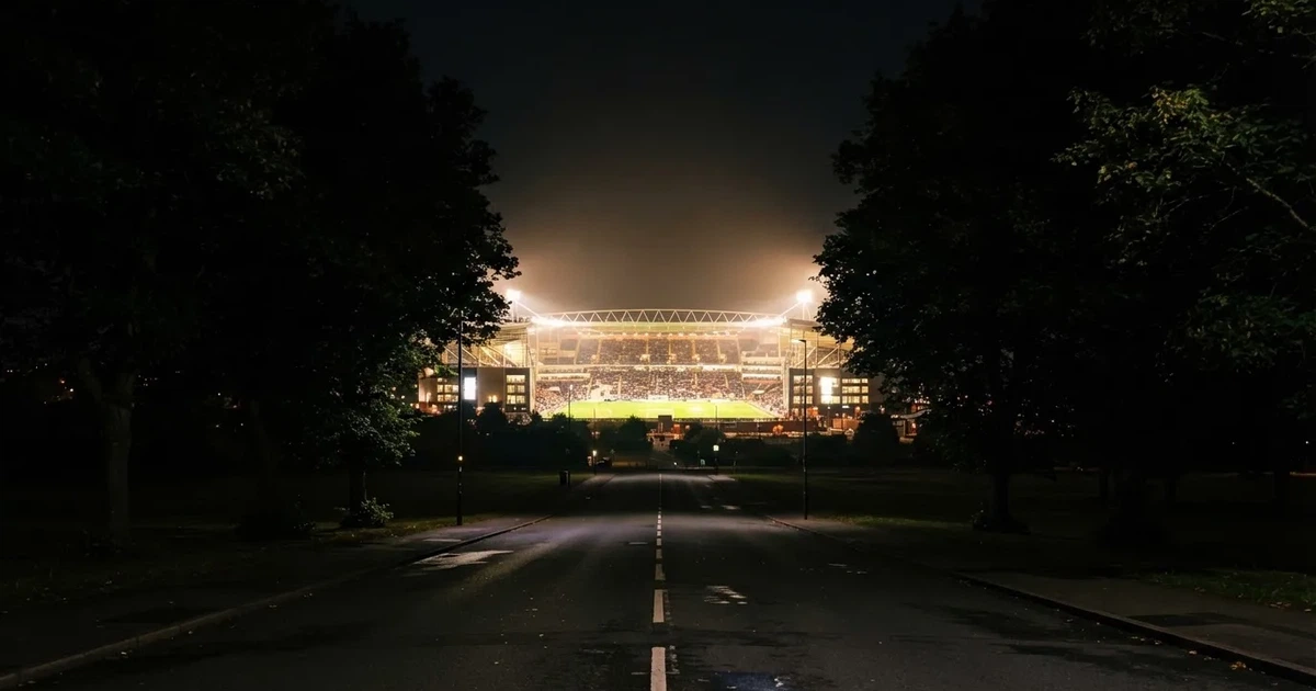 Ein Fußballstadion bei Nacht aus großer Entfernung, hell erleuchtet unter Flutlicht, davor eine dunkle Straße und Bäume.
